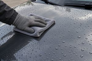A gloved hand wiping the dark bonnet of a car with a grey microfibre cloth, with water beads on the paint.